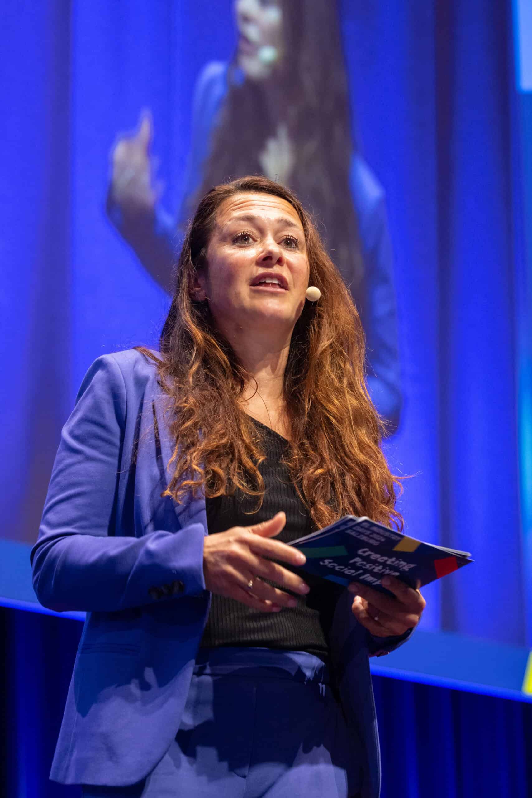 Corporate Sociologist Suzanne Mau-Asam can be seen on stage from the waist up. Wearing a blue blazer and holding cue cards, she is captured as a keynote speaker in the middle of presenting at the European Lottery Industry Days with a video screen of her in the background.